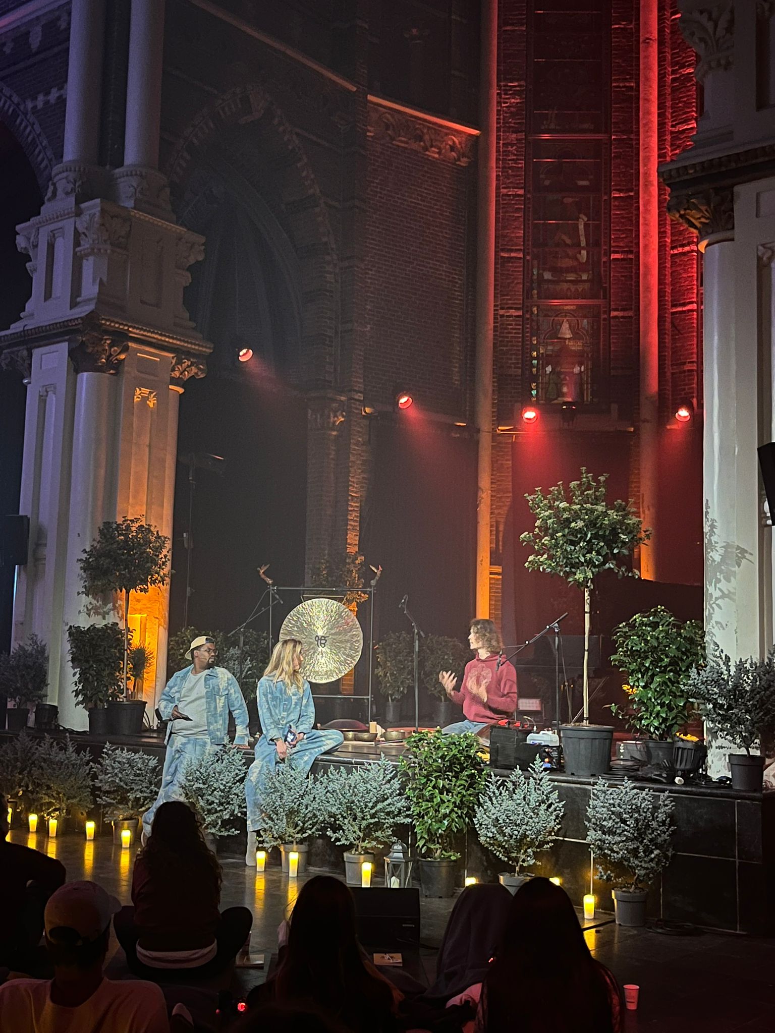 An evening gathering in a candlelit hall, plants and a gong on stage, three people in conversation