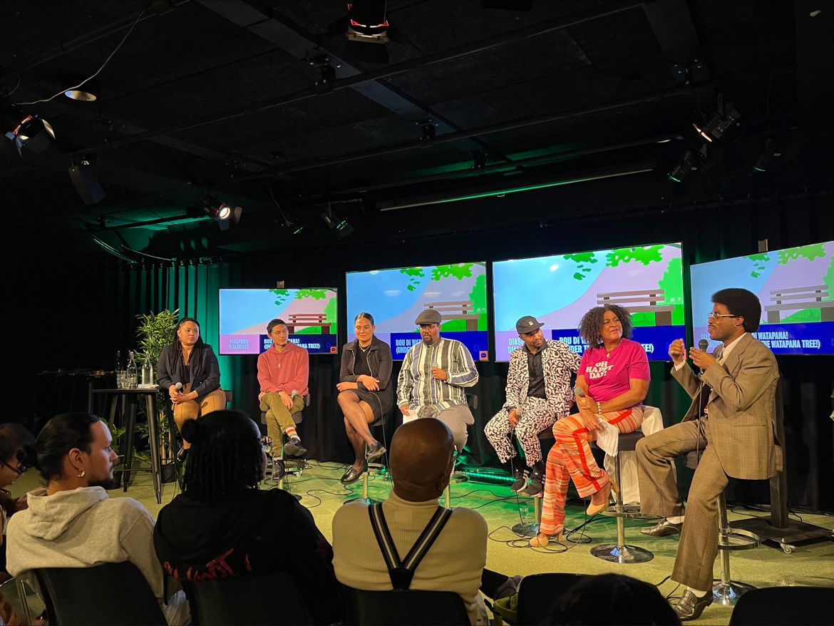 A panel of seven seated on stage at Bou di Watapana, with the series backdrop on screens behind them